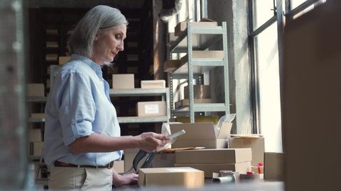 Female mature senior small business owner using mobile app checking parcel box. Warehouse worker, seller holding phone scanning retail dropshipping package postal parcel on cell preparing ship order.