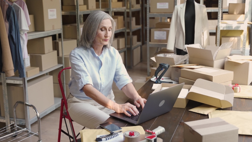 Older female fashion dropshipping business owner using laptop talking on phone in warehouse. Mature woman seller merchant checking online website order confirming ecommerce shipping parcel delivery. Royalty-Free Stock Footage #1060830664
