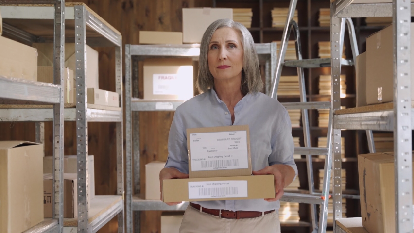 Confident mature middle aged woman retail seller, entrepreneur, clothing store small business owner walking in delivery dropshipping warehouse storage holding parcel boxes preparing ship delivery. Royalty-Free Stock Footage #1060830670