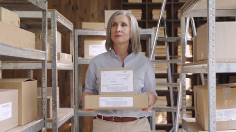 Confident mature middle aged woman retail seller, entrepreneur, clothing store small business owner walking in delivery dropshipping warehouse storage holding parcel boxes preparing ship delivery.