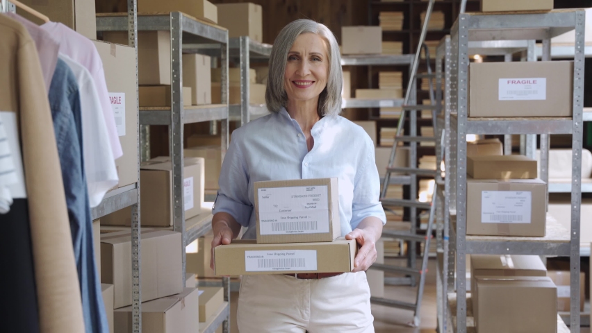 Confident smiling mature older 60s woman retail seller, entrepreneur, clothing store small business owner, looking at camera holding parcel boxes in delivery shipping warehouse storage, portrait. Royalty-Free Stock Footage #1060830673