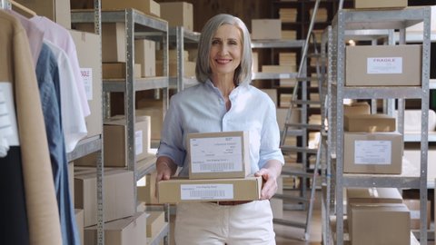Confident smiling mature older 60s woman retail seller, entrepreneur, clothing store small business owner, looking at camera holding parcel boxes in delivery shipping warehouse storage, portrait.