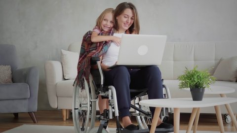 Candid handicapped woman in wheelchair using laptop computer at home, daughter embrace disabled mother, caregiving concept,
