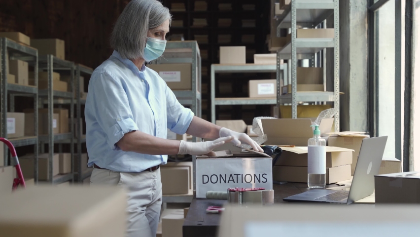 Mature female warehouse worker volunteer wearing face mask working in shipping delivery charitable stock organization packing donations box. Covid 19 coronavirus donating and volunteering concept. Royalty-Free Stock Footage #1060832188
