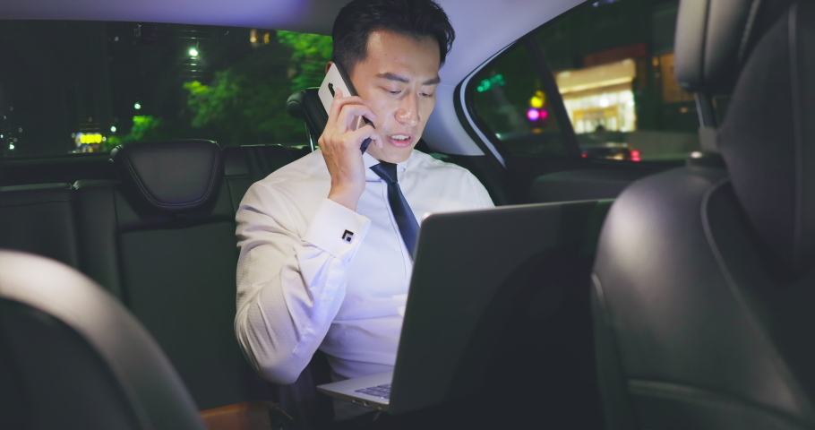 asian businessman speaks on smart phone and uses laptop computer to work while commuting by taxi at night Royalty-Free Stock Footage #1060832371