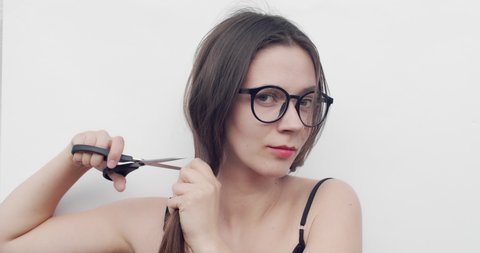 Woman With Glasses Cuts Her Hair On White Background. Young Female Cuts Her Long Brown Hair With Scissors By Herself During Self-isolation. Change Hairstyle For Stress Overcome And Hair Care Concept