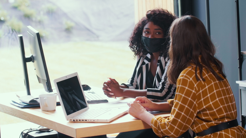 Medical safety at workplace. Two beautiful happy multiethnic business women work together at office wearing face masks. Royalty-Free Stock Footage #1060838485