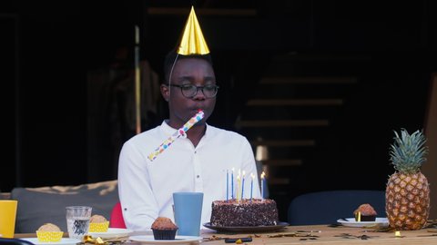 Self isolation. Sad lonely African young handsome man blows whistle alone celebrating his own birthday with cake at home