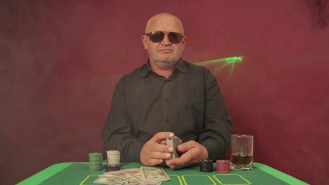 A man in a casino at a gambling table shuffles the cards. Green laser lights and smoke in the background. There are chips and dollars. The player is wearing black glasses. UHD.