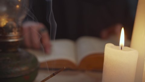 Close-up of burning candle lights and incense sticks standing on table with blurry monk in black robe running his finger under printed words of vintage book, studying ancient history in background.