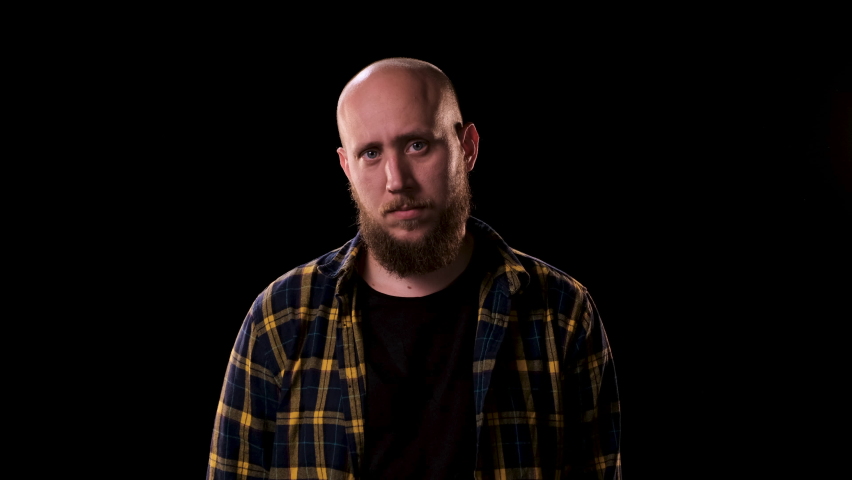 Bald bearded man in checkered shirt looks intently and angrily at the camera on black background. guy smiles and we see that he is missing one front tooth.  | Shutterstock HD Video #1060843660