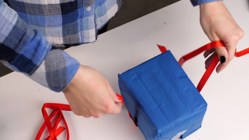 Female well-groomed hands tie a red ribbon around a blue gift box. Correctly or right tying the ribbon around the gift. Christmas or New year. Woman wrapping a surprise birthday present. Festive mood.