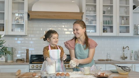 Funny little daughter and young mother have fun making dough for tasty pie at large wooden table in contemporary kitchen