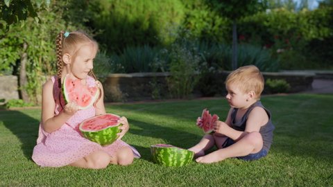 children bite a slice of watermelon outdoors, boy and girl eat fruit while sitting in the park on green lawn