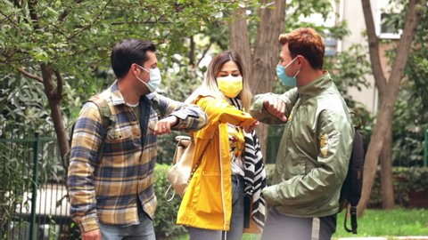 Group of young people in the park chatting and touching elbows instead of high-five or hand shaking. Three good looking friends in the times of Coronavirus having fun and wearing medical mask. 