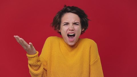 Angry mad young woman 20s years old in yellow sweater posing isolated on red background in studio. People sincere emotions lifestyle concept. Looking at camera screaming scolding protest waving hands