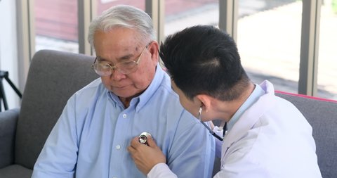 Asian doctor listen heart rate by stethoscope to senior elderly patient in living room at home.
