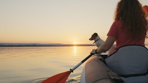 Happy young woman paddling on an inflatable kayak with her dog Jack Russell Terrier on water of sea against beautiful orange sunset slow motion. Family Sports. Summer. Travel. Tourism. Relax. Freedom