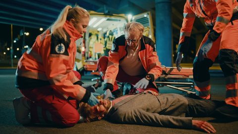 Team of EMS Paramedics Bring a Stretcher from Ambulance Vehicle and Help an Injured Young Person. Emergency Care Assistants Arrived on the Scene of a Traffic Accident on a Street at Night.