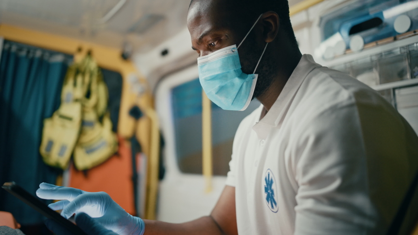 Black African American Paramedic in Face Mask Using Tablet Computer while Riding in an Ambulance Vehicle for an Emergency. Emergency Medical Technicians Outside the Healthcare Hospital. Royalty-Free Stock Footage #1060859485