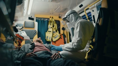 EMS Paramedic in Disposable Coverall Suit Comforting Injured Patient on the Way to Hospital. Emergency Medical Care Assistant Puts His Hand on Vinctim's Shoulder in a Friendly Way in an Ambulance.