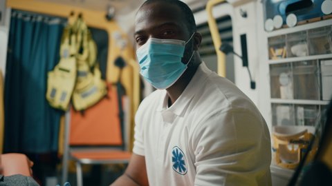 Calm Black African American EMS Professional Paramedic Looks at Camera While Wearing a Safety Face Mask in Ambulance Vehicle. Successful Emergency Medical Technician Outside the Healthcare Hospital.