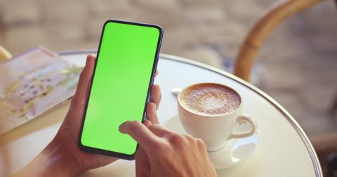 Crop view of female person holding smartphone and scrolling on green screen while sitting at table with coffee and map on it. Concept of mockup and chroma key