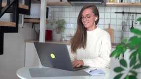 Young happy smiling woman wearing round glasses and headphones makes a note in the notebook during working online at a laptop at home 