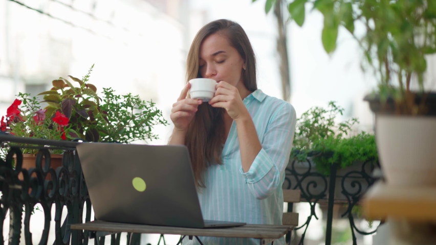 Beautiful happy cute joyful smiling romantic woman blogger drinking an aromatic coffee while working online at a laptop on a balcony | Shutterstock HD Video #1060863301