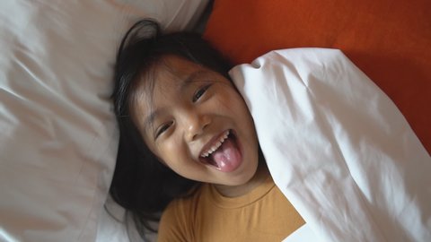 Asian child cheerful or kid girl sleeping and wake up with play peekaboo in blanket by father or parents for relax on white bed in bedroom and smile laugh for enjoy happy fun in morning on holiday