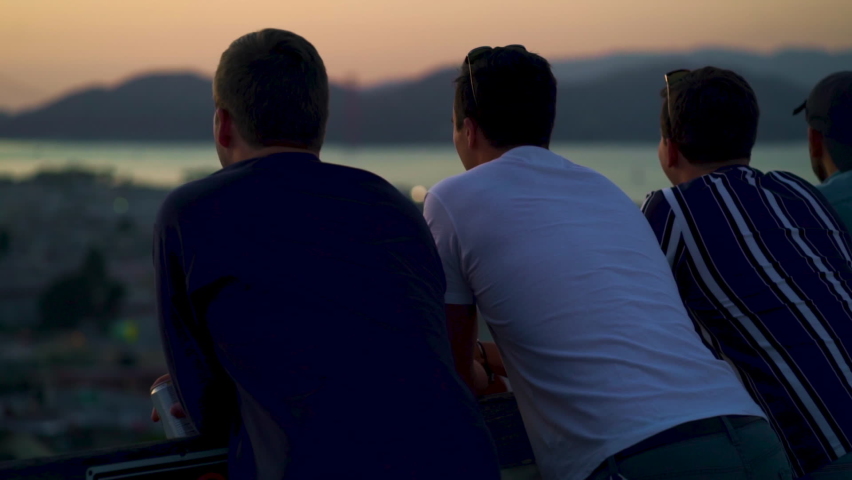 Three men on the roof, enjoying the sunset and looking over the San Francisco cityscape and the Golden gate bridge, during dusk, in California, USA - Pull focus, pan shot