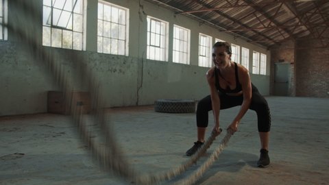Fit female athlete moving a rope in wave motion as part of fitness regime. Tough woman working out with battling rope in cross training workout space.
