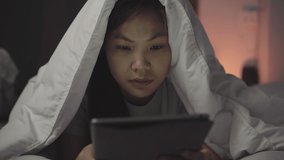 The woman lying in bed yawning while using the touchpad ,hiding head under blanket then went to bed.