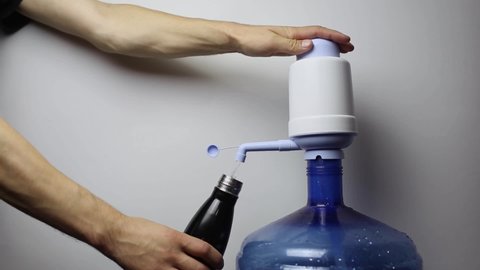 Close-up of male hands pouring water into the steel thermo bottle, from big plastic bottle with pump dispenser in office.