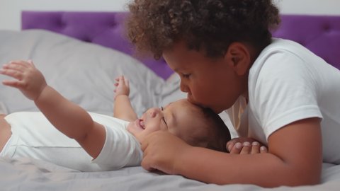 African preschool boy kissing newborn brother lying together on bed at home. Portrait of adorable baby and toddler boy siblings playing and relaxing. Childhood and family concept