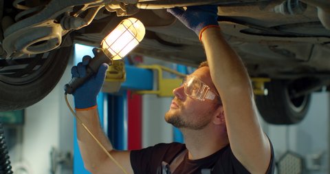 Car service - mechanic check out automobile parts while working with lamp. Auto mechanic working underneath car lifting machine at the garage.