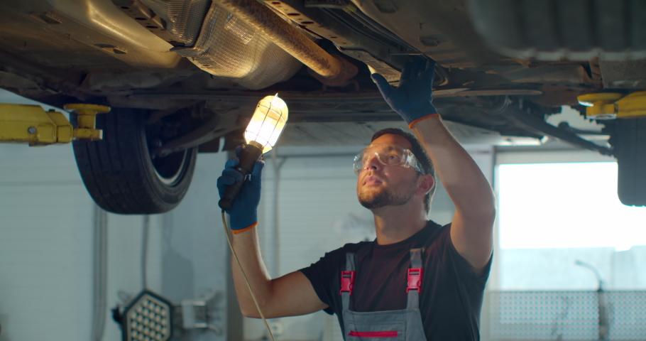 Car service - mechanic check out automobile parts while working with lamp. Auto mechanic working underneath car lifting machine at the garage. Royalty-Free Stock Footage #1060877257