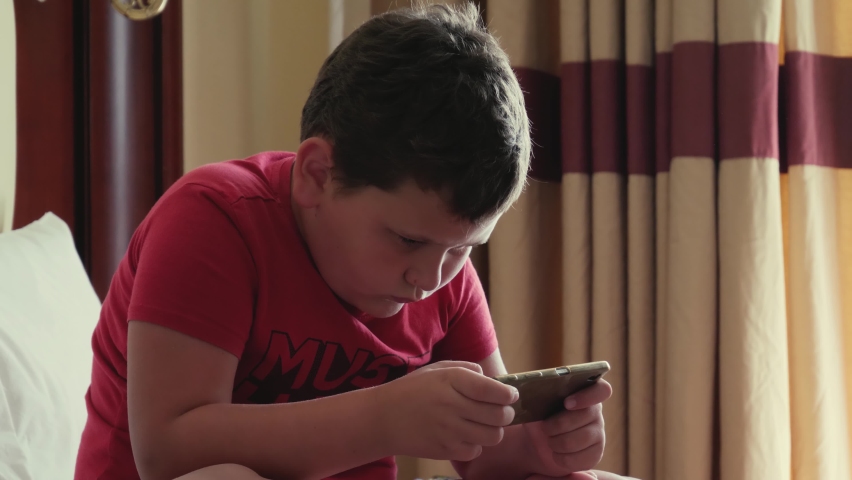 Plump little boy in a red T-shirt on the bed uses the phone, spends time playing on the net or studying while staying at home