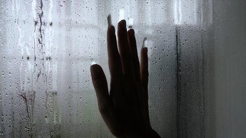 Close up silhouette of palm of woman hand touching window and slides down glass misted with drops, leaving trace. Hand of hope and despair in period of self isolation of pandemic coronavirus covid-19