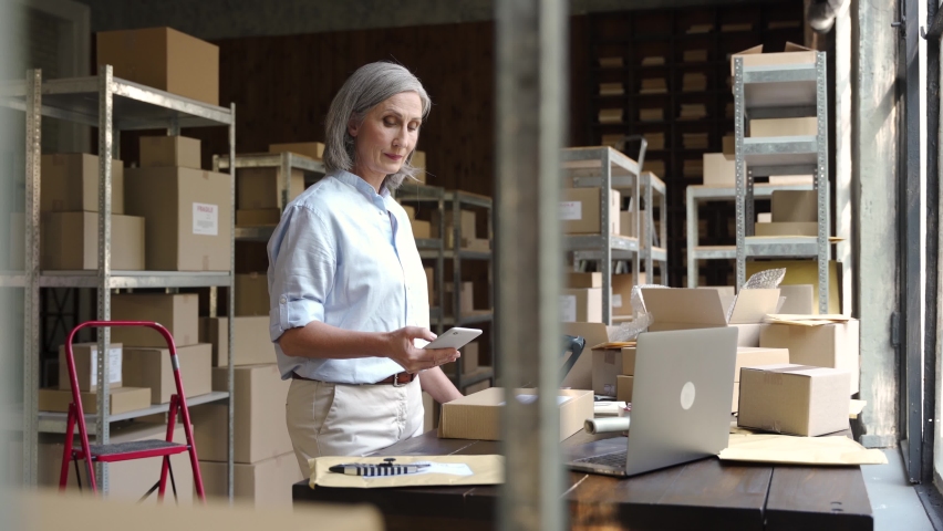 Older female online store ecommerce small business owner, warehouse worker, merchant seller packing shipping package using mobile app preparing delivery parcel box. Dropshipping distribution. | Shutterstock HD Video #1060886395
