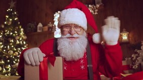Happy Santa Claus, Saint Nicholas waving hand holding present talking to camera video calling kid or recording Merry Christmas virtual greetings on xmas eve sitting at table at home, webcam view. - Powered by Shutterstock - Get 15% off with code: PIKWIZARD15