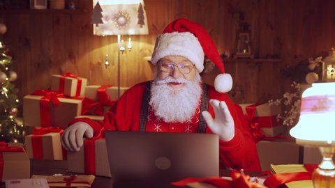 Happy Santa Claus holding gift box video calling kid talking to child in virtual video online chat meeting on laptop sit at home table late with present on xmas eve. Merry Christmas social distance.