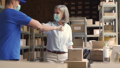 Warehouse workers wears masks gives elbow bump at meeting. Female supervisor greets male courier deliverer takes shipping parcels delivery boxes. Covid 19 healthcare protection social distance at work