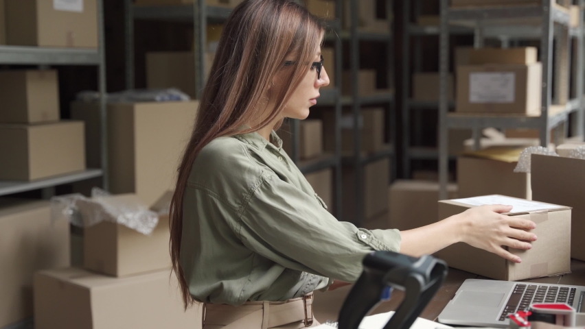 Young female seller or online store worker scanning parcel packing ecommerce post shipping box checking online retail store orders fulfillment using laptop in dropshipping delivery service warehouse. Royalty-Free Stock Footage #1060892455