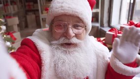Happy old bearded Santa Claus wearing costume and hat holding phone waving hand video calling, recording video Merry Christmas greeting or shooting vlog standing in workshop, face camera view.