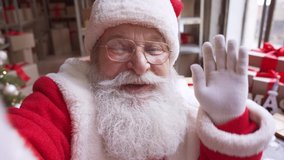 Happy old bearded Santa Claus wearing costume and hat holding phone waving hand video calling, recording video Merry Christmas greeting or shooting vlog standing in workshop, face camera view. - Powered by Shutterstock - Get 15% off with code: PIKWIZARD15