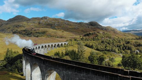 Aerial shot of a train crossing a beautiful bridge in a Scottish hill through a sunny landscape.