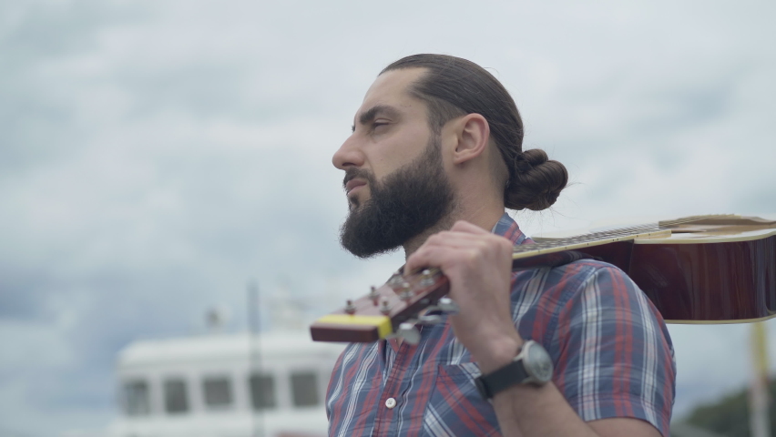 Close-up of bearded handsome man with guitar looking away and thinking. Young Caucasian musician standing with stringed musical instrument outdoors with cloudy sky at the background.