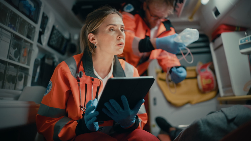 Female EMS Professional Paramedic Using Tablet Computer to Fill a Questionnaire for the Injured Patient on the Way to Hospital. Emergency Care Assistant Comforting the Patient in an Ambulance. Royalty-Free Stock Footage #1060901200