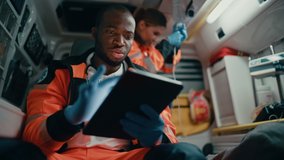 Black African American EMS Professional Paramedic Using Tablet Computer to Fill a Questionnaire for the Injured Patient on the Way to Hospital. Emergency Medical Care Assistant Works in an Ambulance. - Powered by Shutterstock - Get 15% off with code: PIKWIZARD15